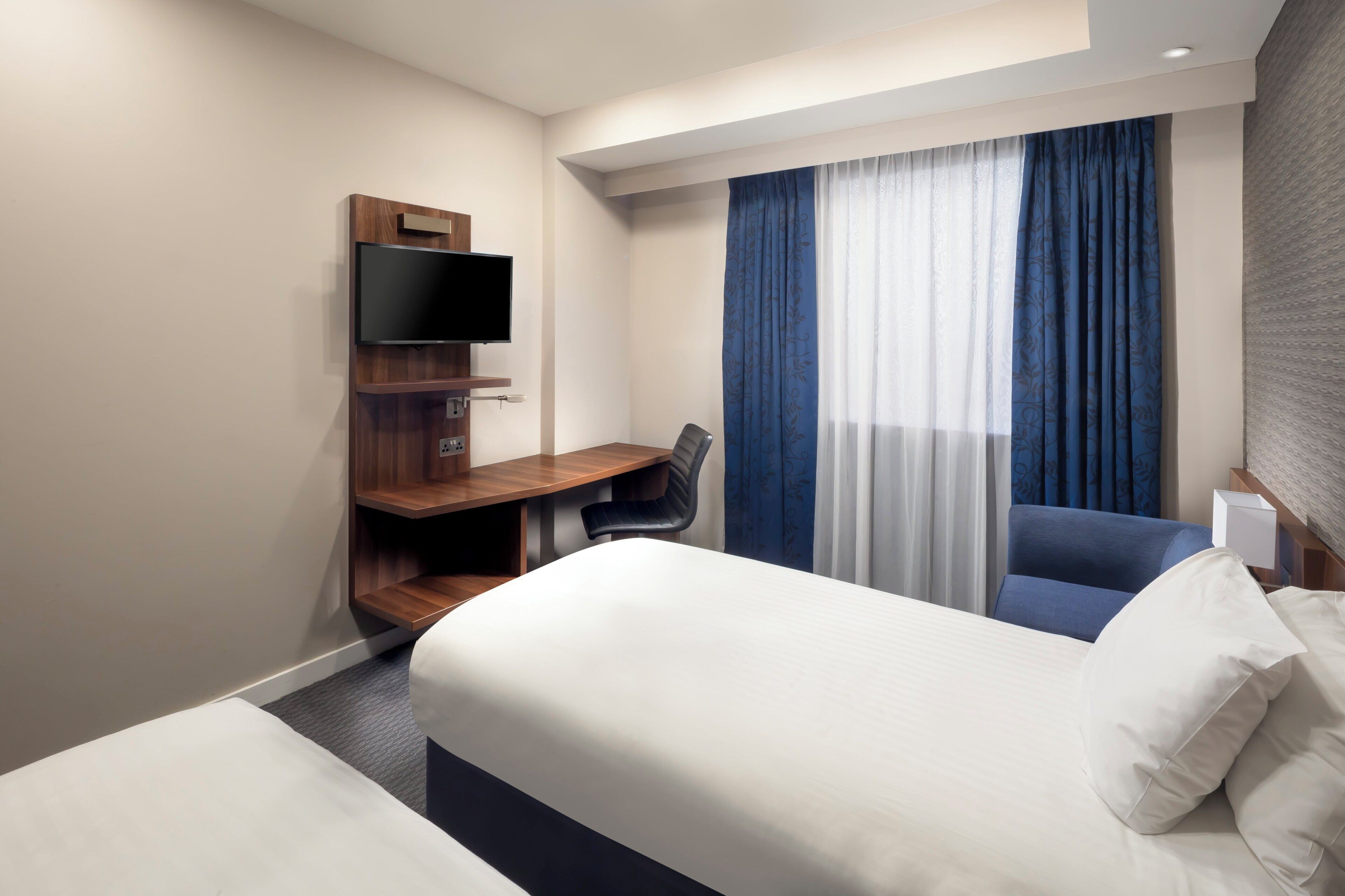A hotel room with a bed, a desk with a flat-screen TV, a chair, and a window with curtains.