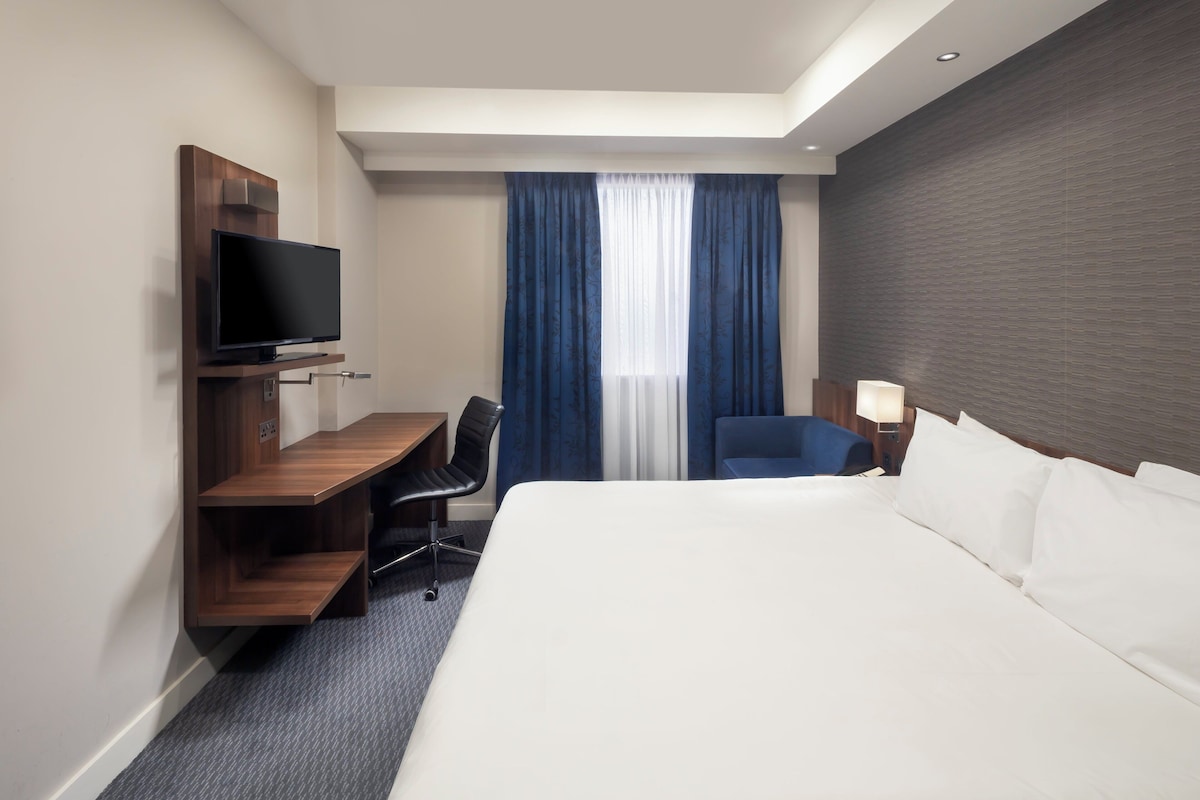 A hotel room with a bed, a desk with a flat-screen TV, a chair, a lamp, and a window with curtains.