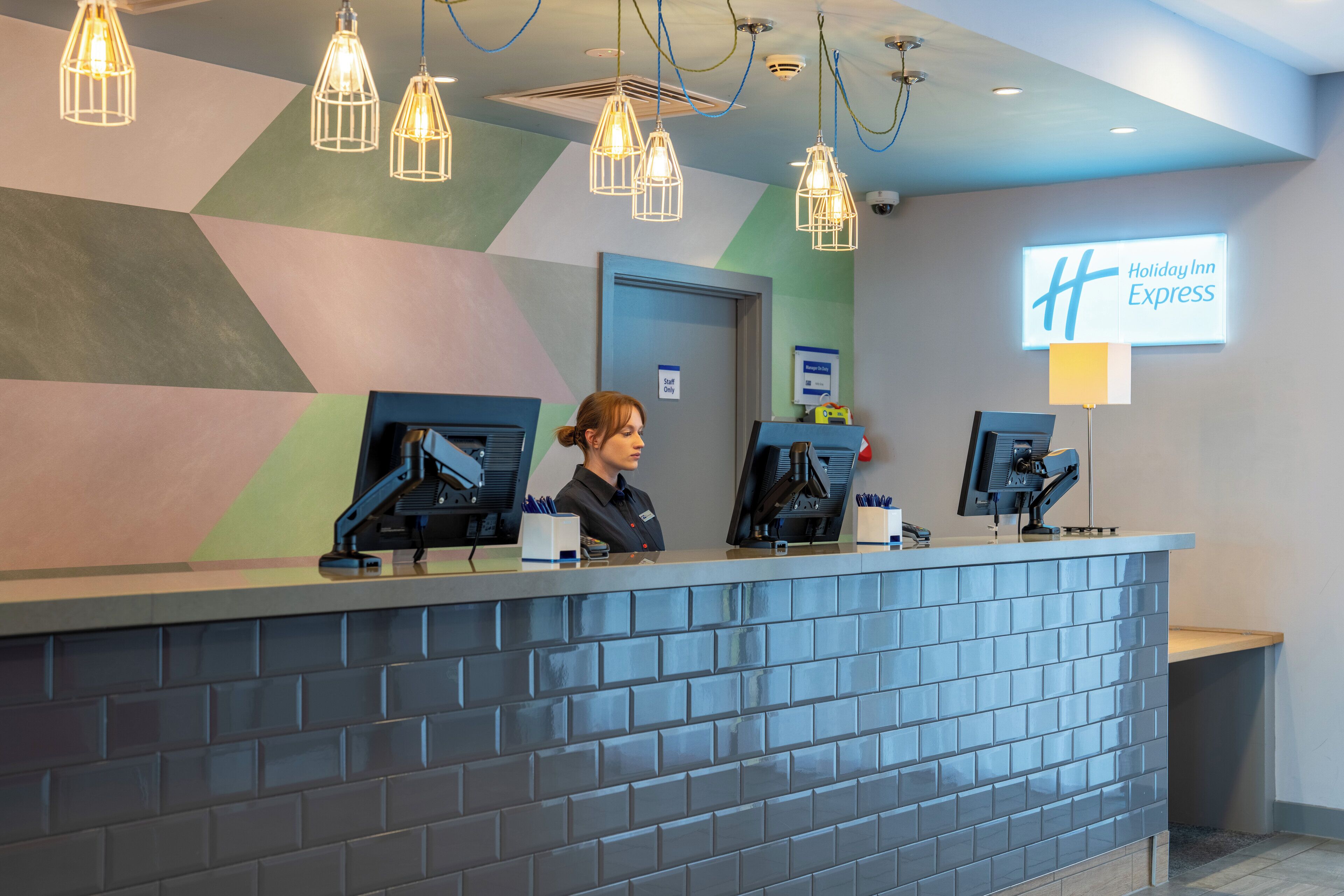 A Holiday Inn Express reception desk with two monitors, a digital sign, and a modern geometric wall design.