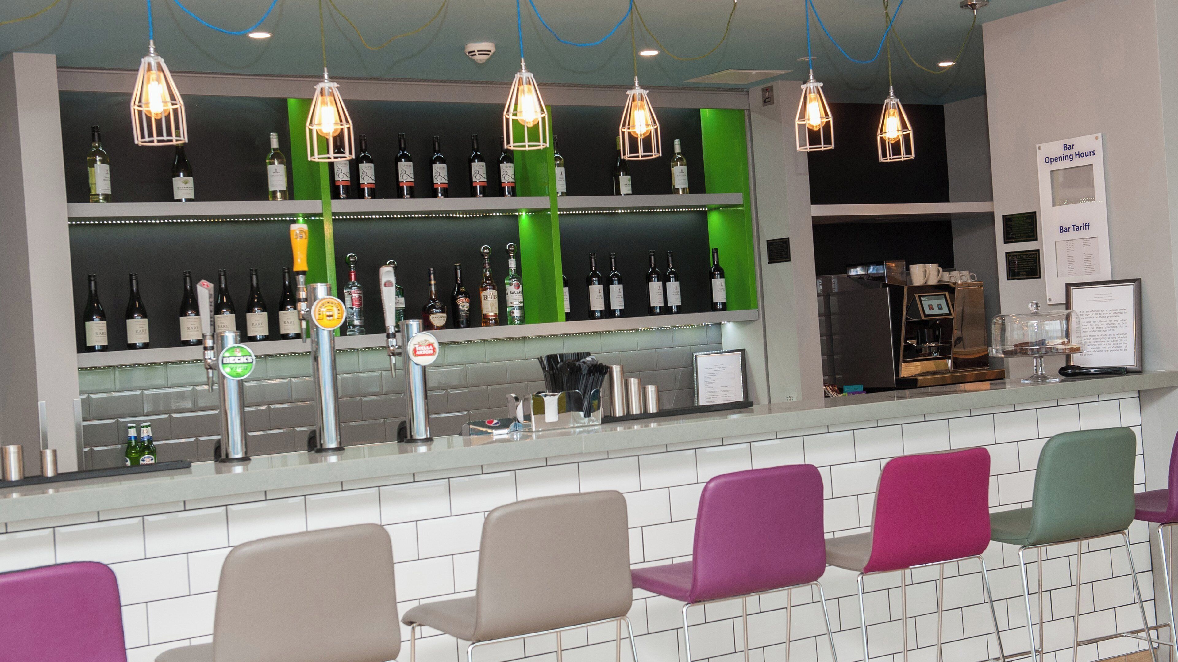 Zona de bar con diversas opciones de asientos, una variedad de botellas en estanterías y un cartel que indica el horario de apertura.