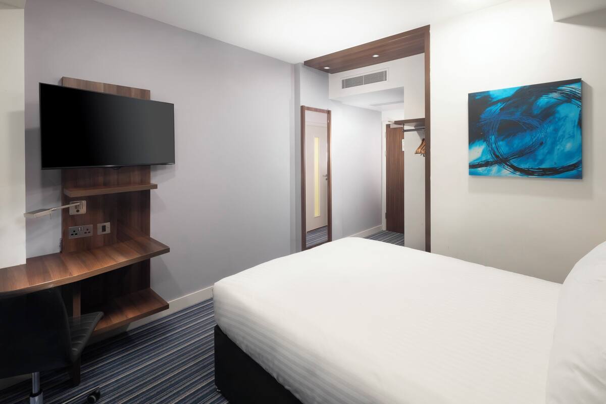 A hotel room with a flat-screen TV mounted on a wooden shelf, a bed with white bedding, a blue abstract painting on the wall, and a door leading to another room.