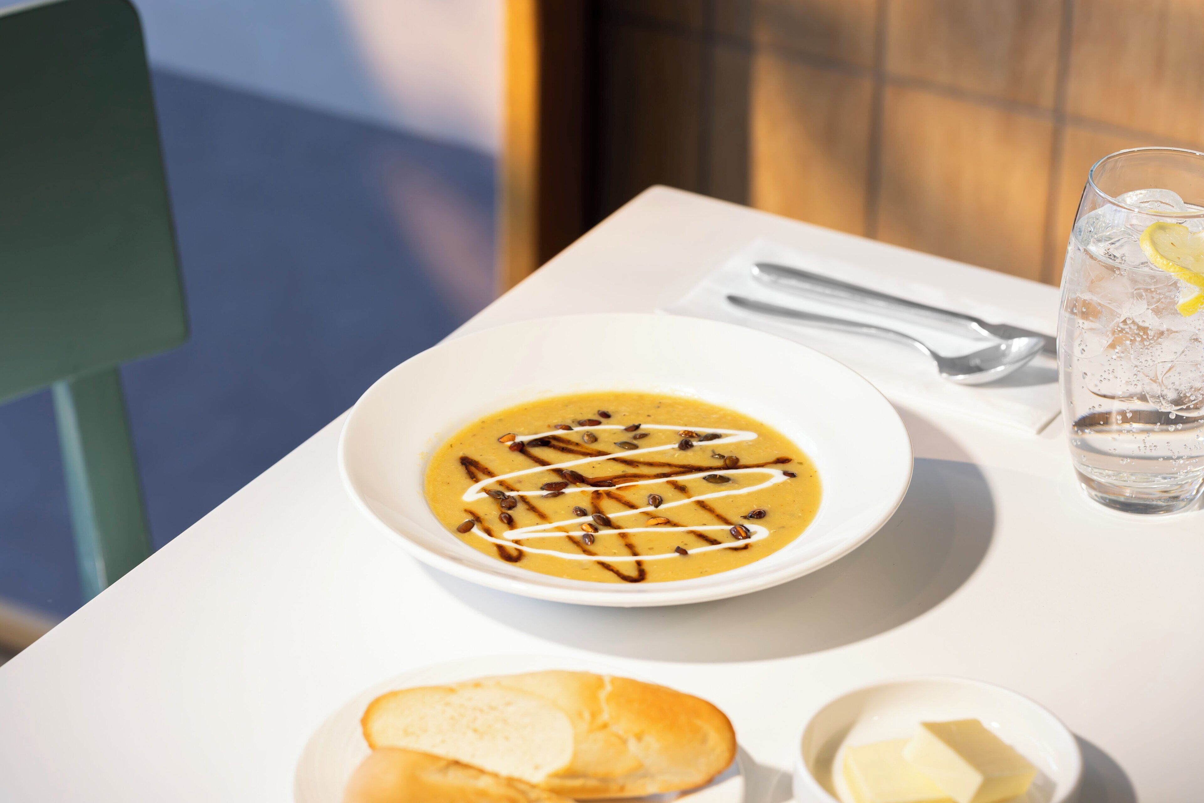 A white table with a bowl of soup, a glass of water, a small plate with butter, and a piece of bread.