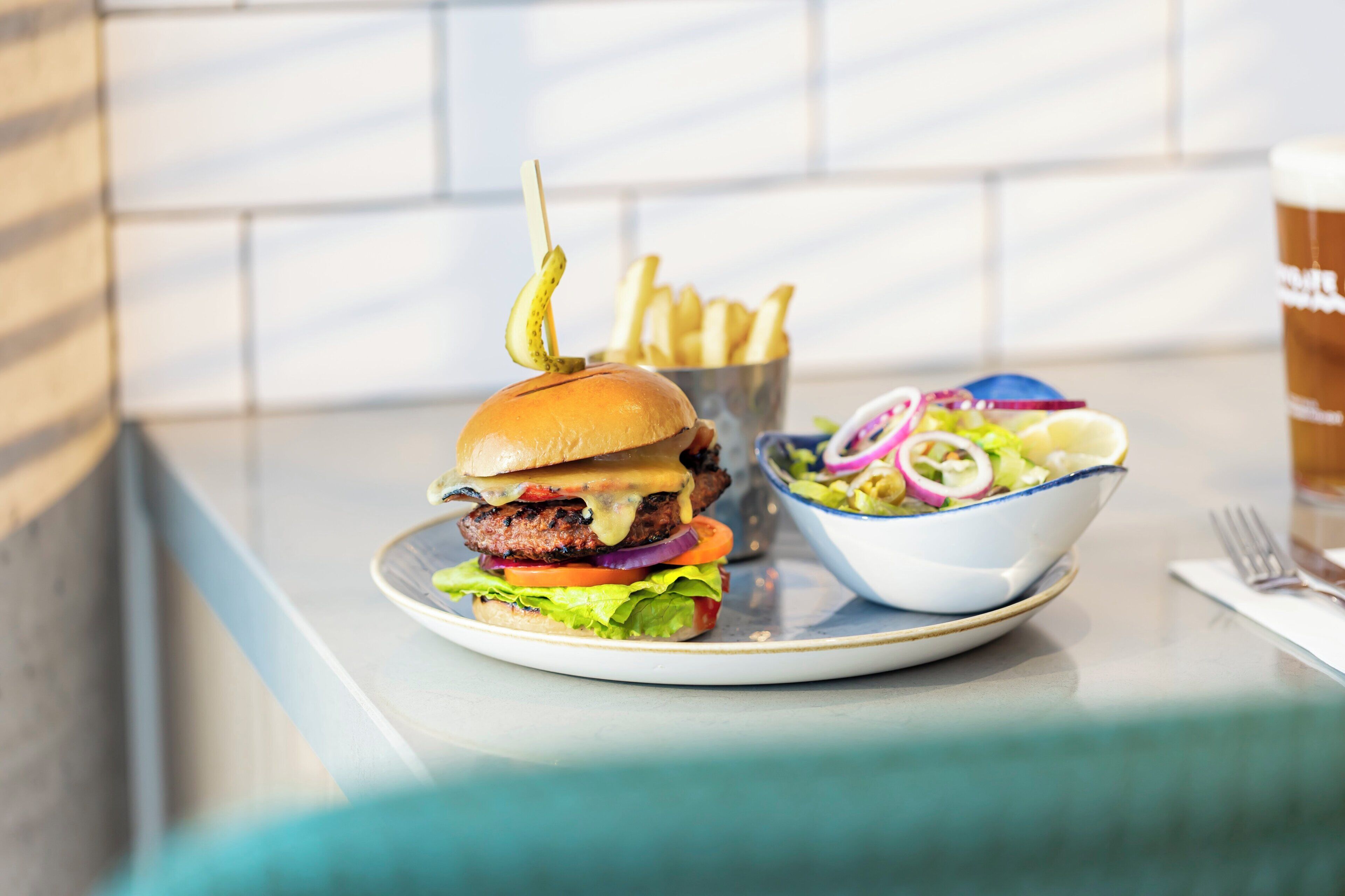 Hamburguesa con papas fritas y ensalada en un plato, y una bebida al fondo.