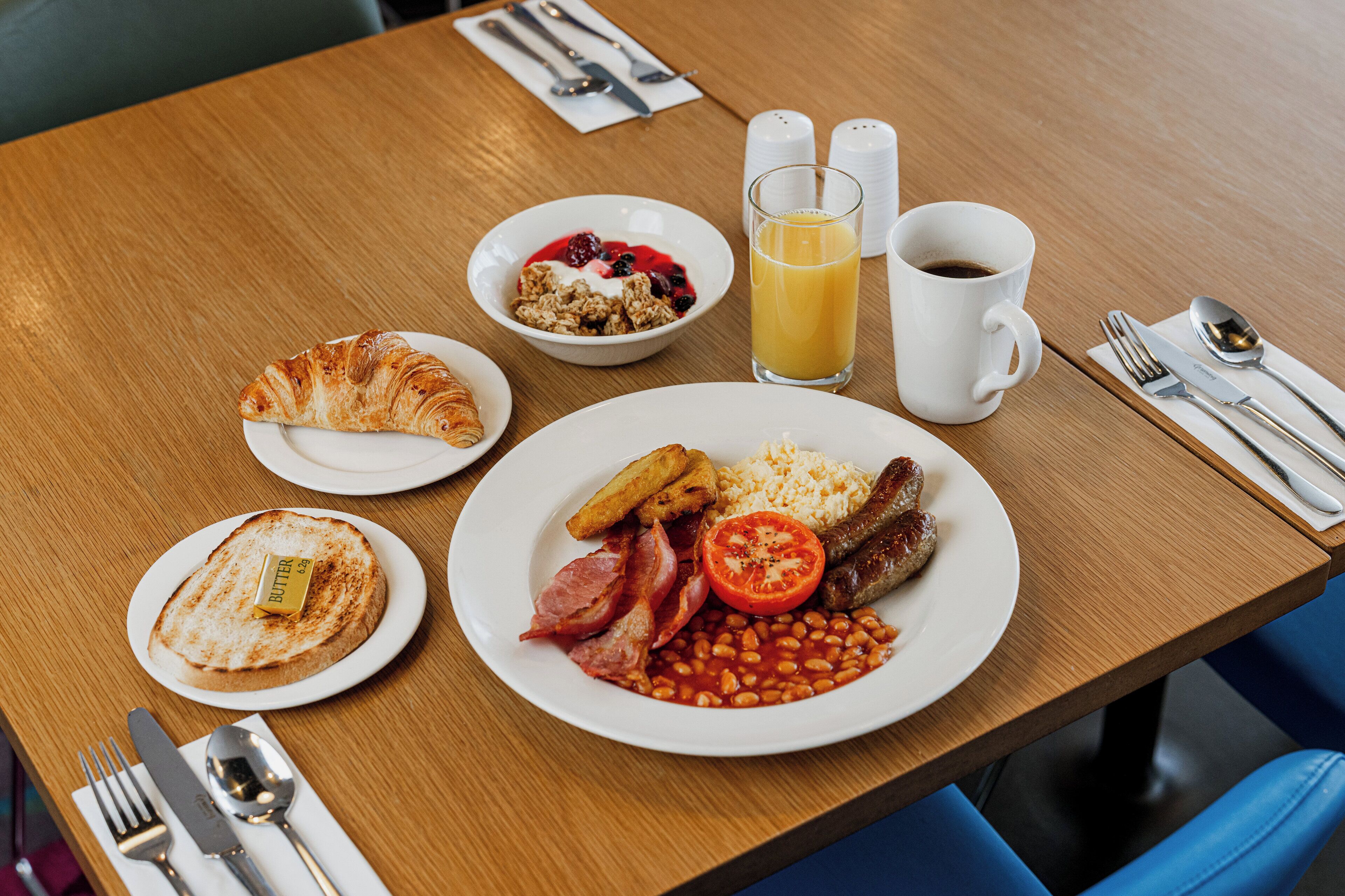A breakfast table with plates of toast, cereal, beans, tomatoes, and sausages.