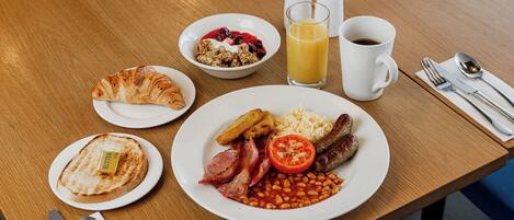 A breakfast table with plates of toast, cereal, beans, tomatoes, and sausages.