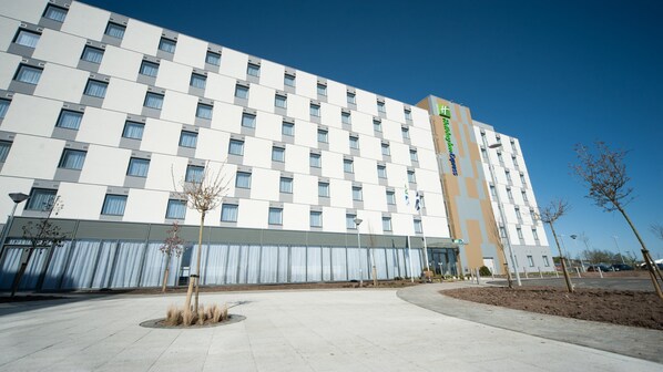 Ein modernes Hotelgebäude mit Schachbrettmuster-Fassade, einer auffälligen grün-weißen Signage und einem begrünten Eingangsbereich mit Bäumen und einer Bank.