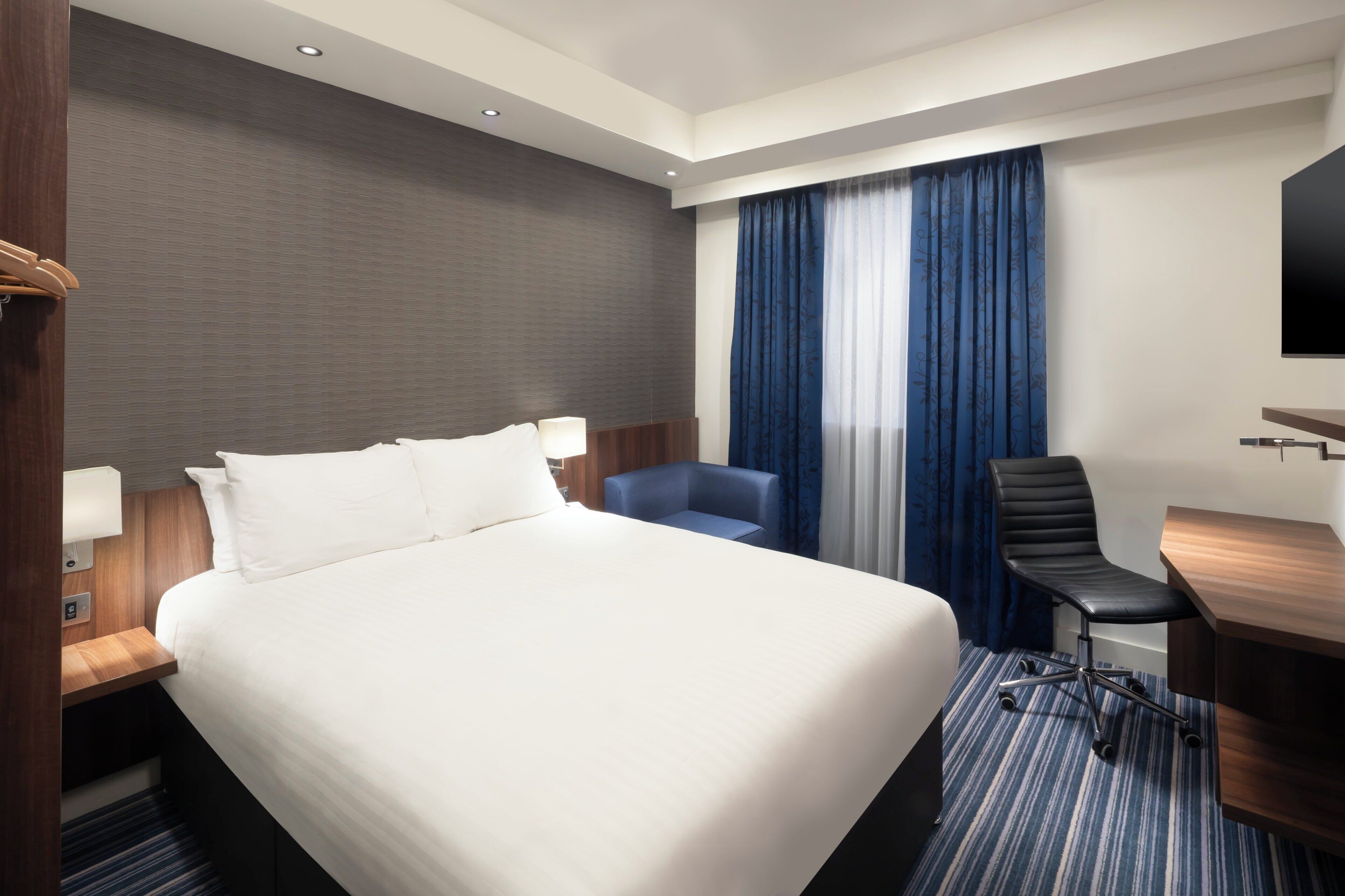A hotel room with a large bed, a desk, a chair, a TV, and a window with curtains.