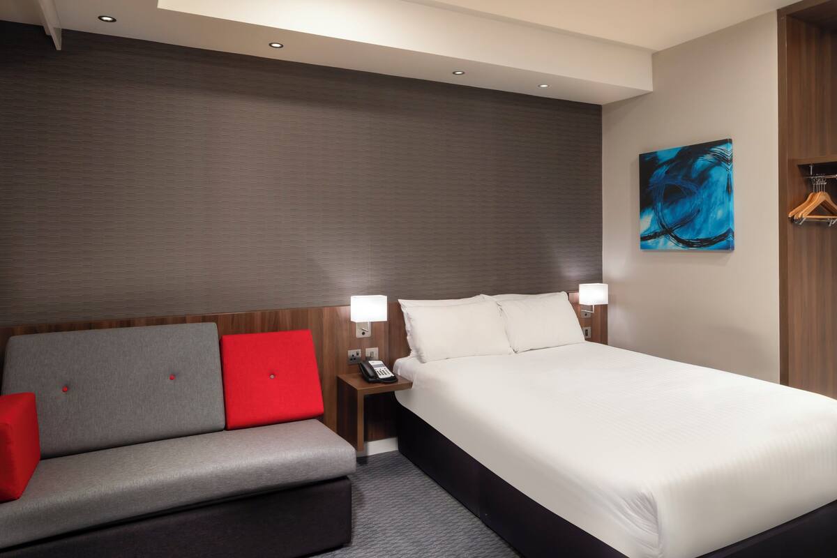 A hotel room with a bed, a sofa with red cushions, a nightstand with a phone, and a painting on the wall.