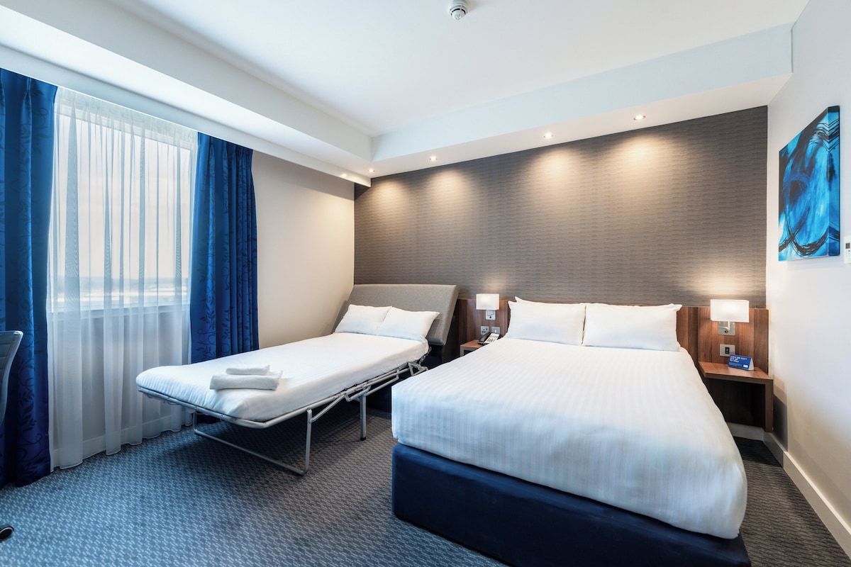 A hotel room with two beds, a large window with blue curtains, a painting on the wall, and a nightstand with a lamp.