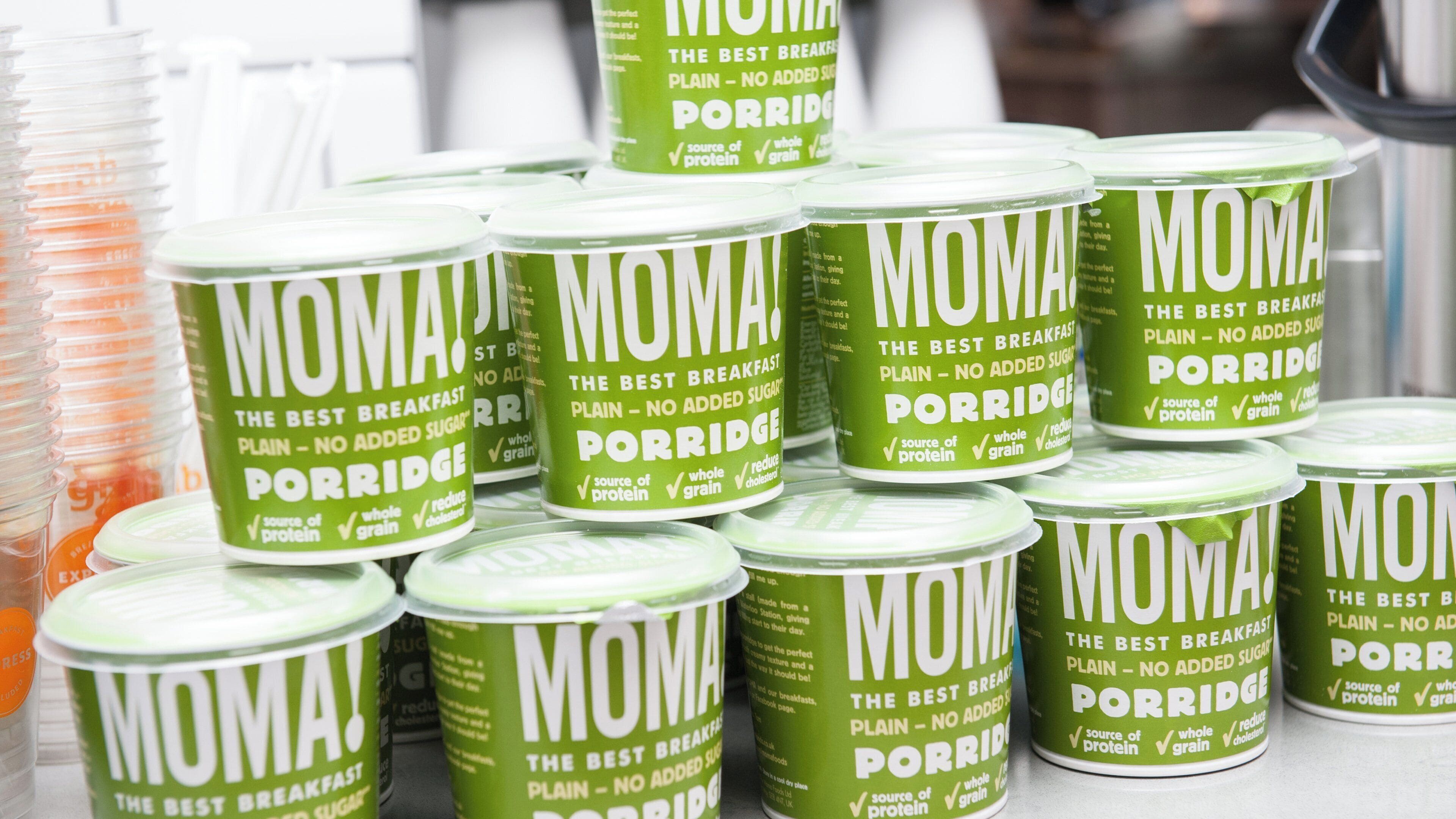 A display of green containers labeled 'MOMA Porridge' with claims of being the best breakfast, plain, no added sugar, source of protein, whole grain, and cholesterol reduction.