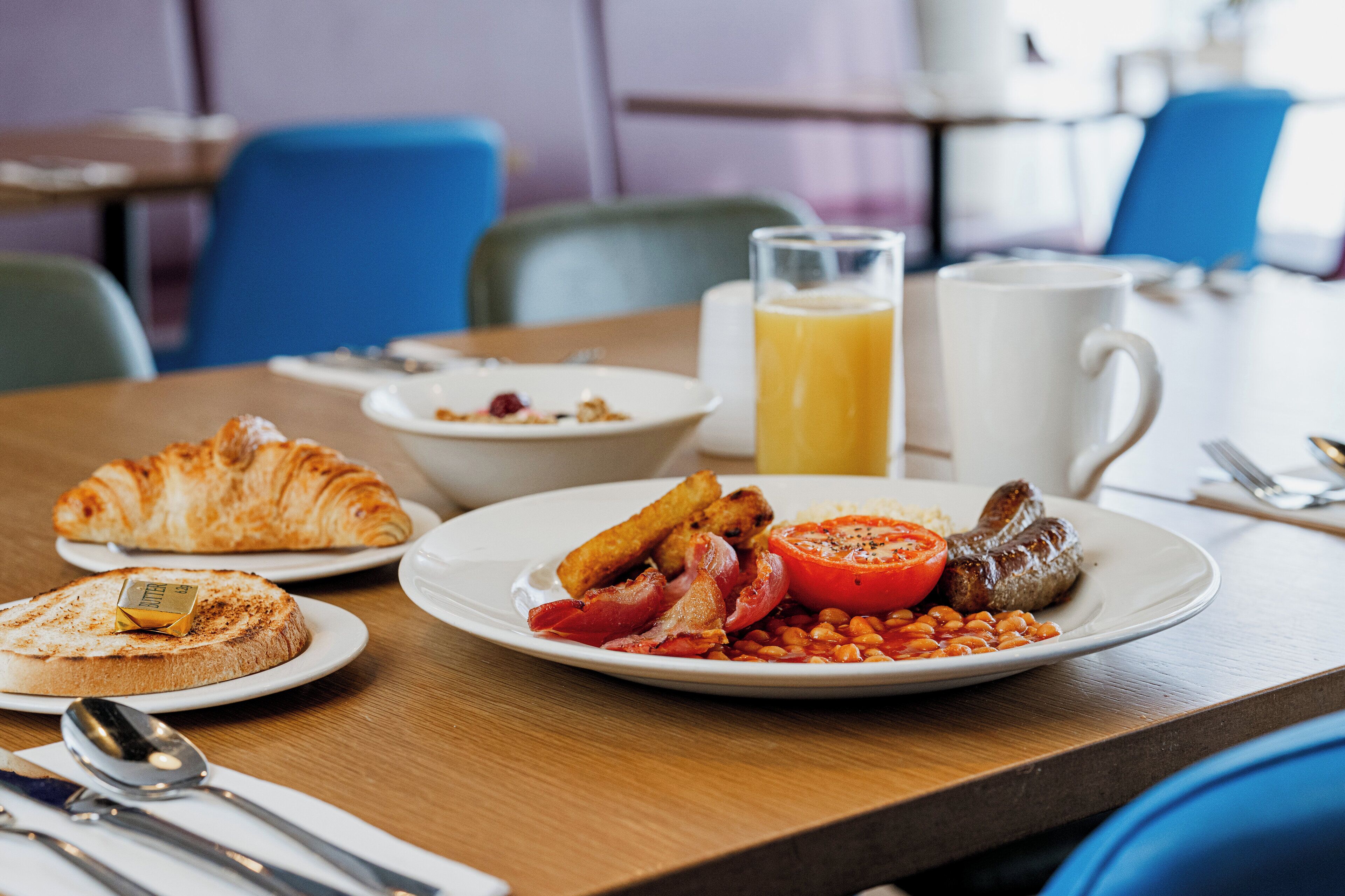 A breakfast table with croissants, toast, beans, bacon, sausages, and a glass of orange juice.
