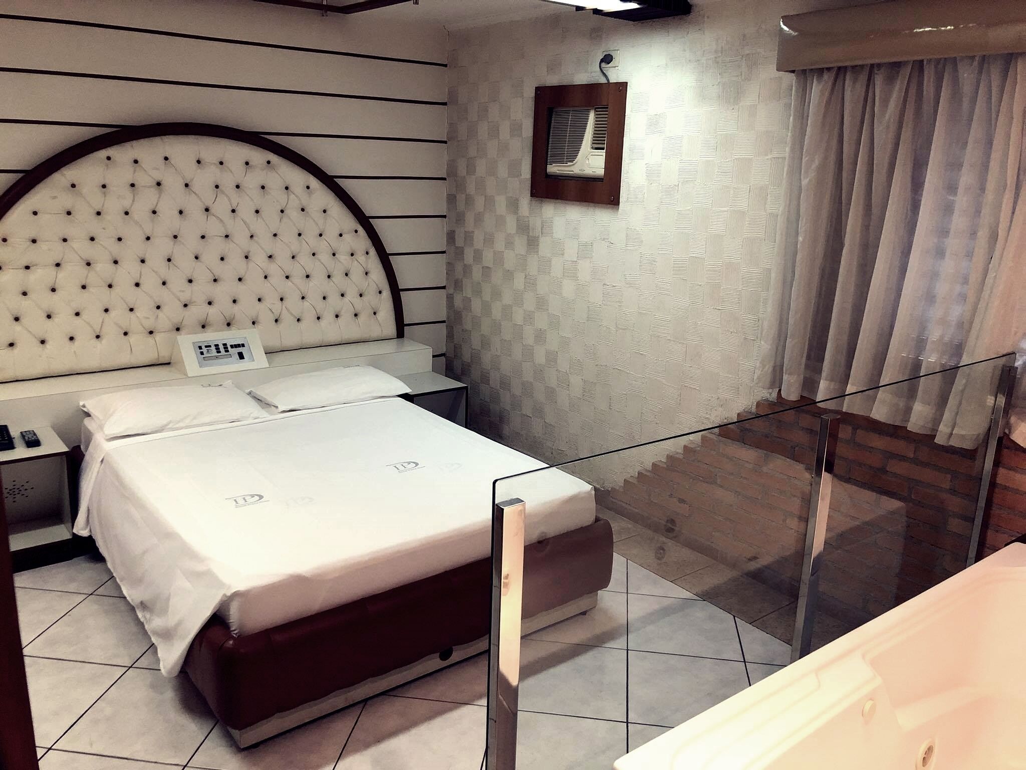 Habitación de hotel con una cama grande, una mesita de noche, un aire acondicionado instalado en la pared y vistas a un muro de ladrillo a través de una partición de vidrio.