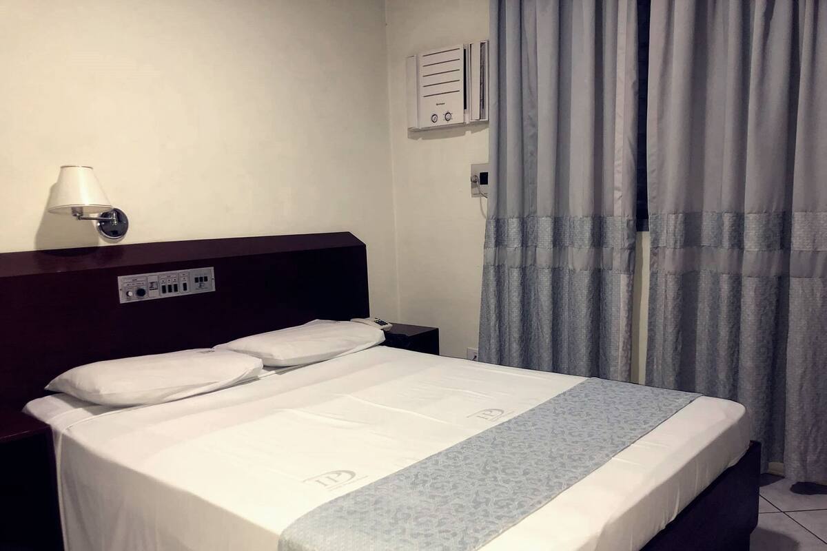 Ein Hotelzimmer mit Bett, Nachttisch, Lampe, Steckdose und einem Fenster mit Vorhängen.
