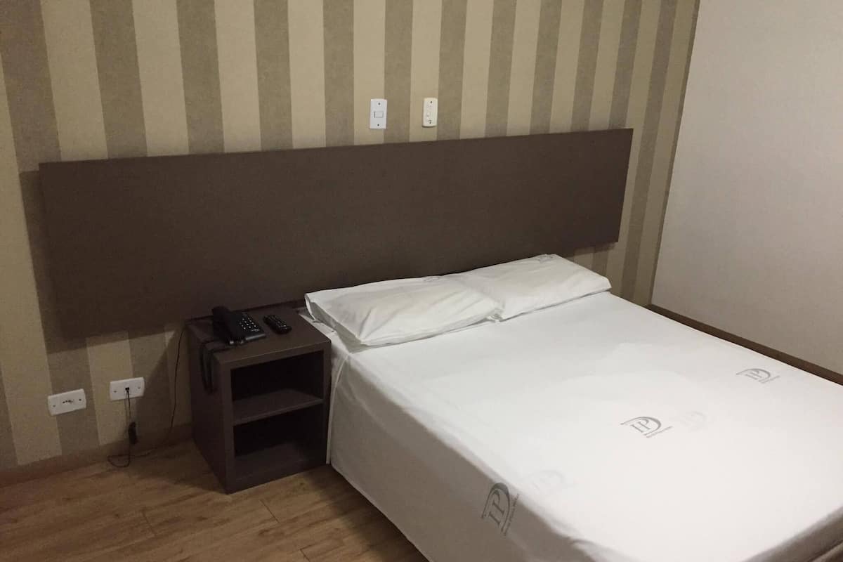 Ein Einzelbett mit weißer Bettwäsche, ein Nachttisch aus Holz mit Telefon und Fernbedienungen sowie eine gestreifte Wand.
