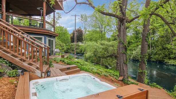 A hot tub on a wooden deck with a river view, surrounded by greenery and a wooden staircase. - Stone Waters Inn (Bellaire)