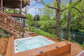 A hot tub on a wooden deck with a river view, surrounded by greenery and a wooden staircase.