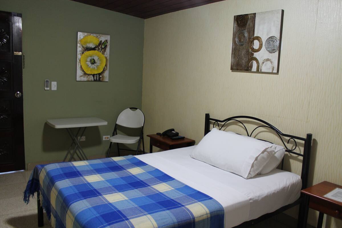 A bedroom with a bed, a chair, a small table, and two paintings on the wall.
