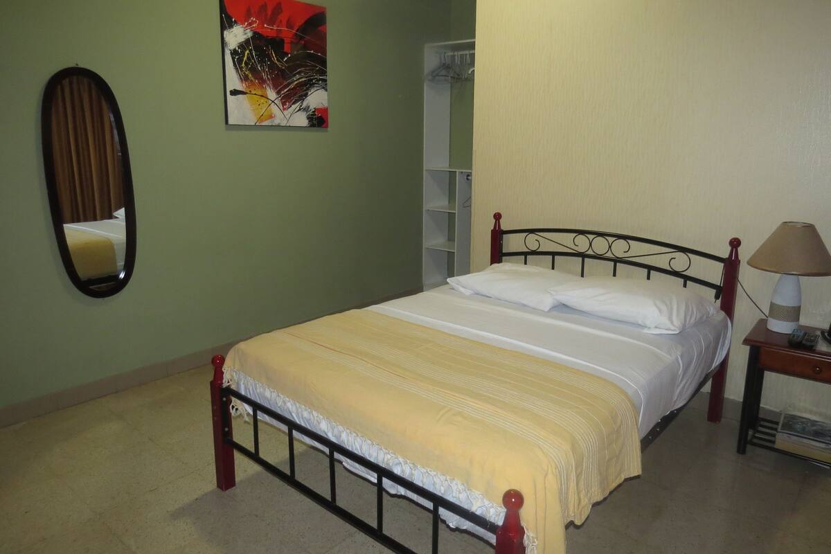 A neatly made bed with a white and yellow bedspread, a red headboard, and a bedside table with a lamp.