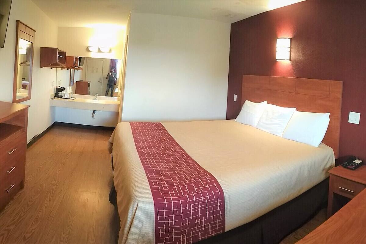 A hotel room with a bed, a desk, a chair, a microwave, a refrigerator, a mirror, and a television.