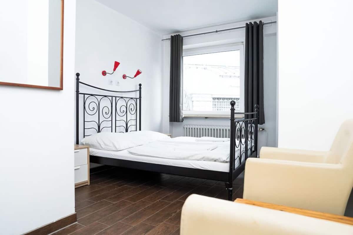 Ein Schlafzimmer mit Bett, Nachttisch, Fenster mit Vorhängen und einem Sessel.
