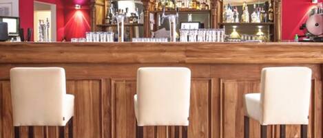 A bar area with a wooden counter, three bar stools, and a variety of bottles and glasses on shelves.