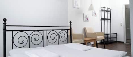 Una cama bien hecha con sábanas blancas, cabecera de metal negro, un sillón color beige y una mesita de noche con una lámpara.