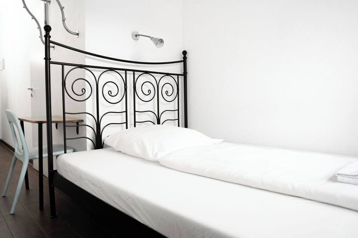 Ein Einzelbett mit Metallgestell und weißer Bettwäsche, ein kleiner Tisch mit Stuhl sowie eine Wandleuchte.