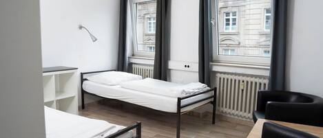 A small, modern room with a single bed, a desk, and a chair. There are large windows with curtains, a radiator, and a lamp.