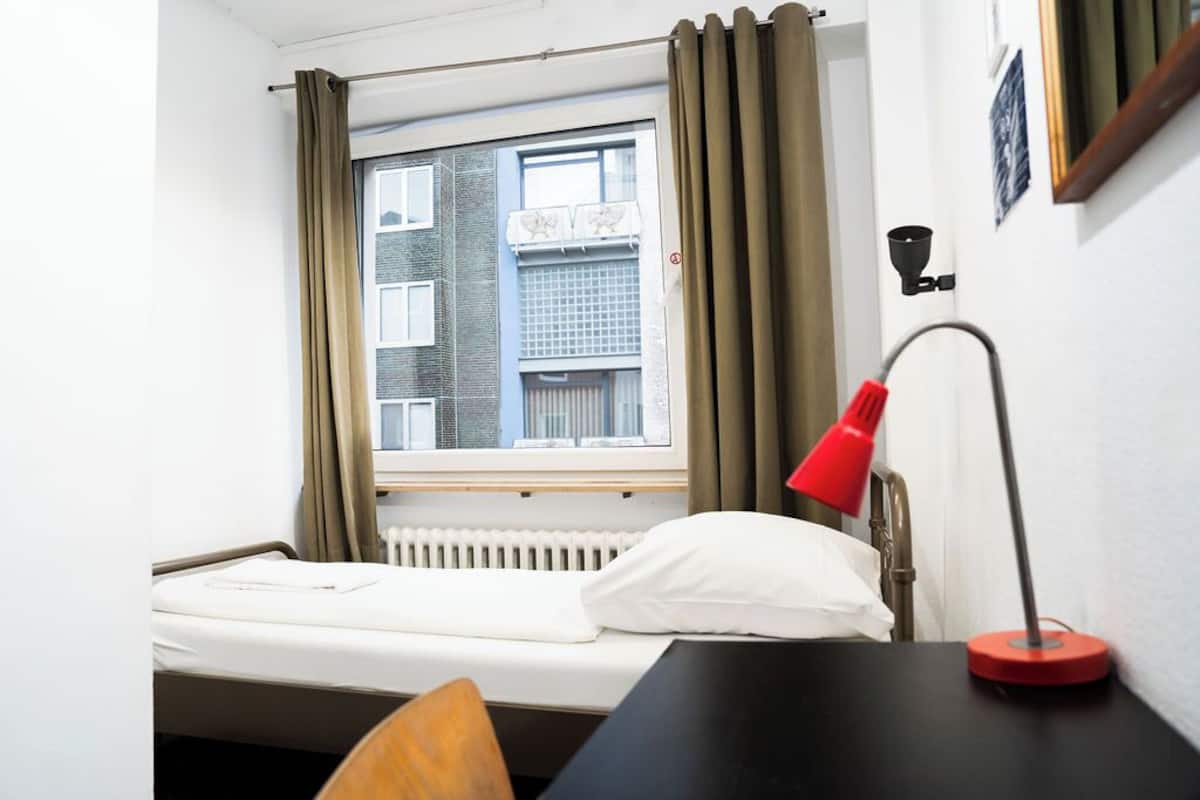 Ein kleines Zimmer mit einem Einzelbett, einem Fenster, einer Heizung, einem Schreibtisch mit Lampe und einem Stuhl.