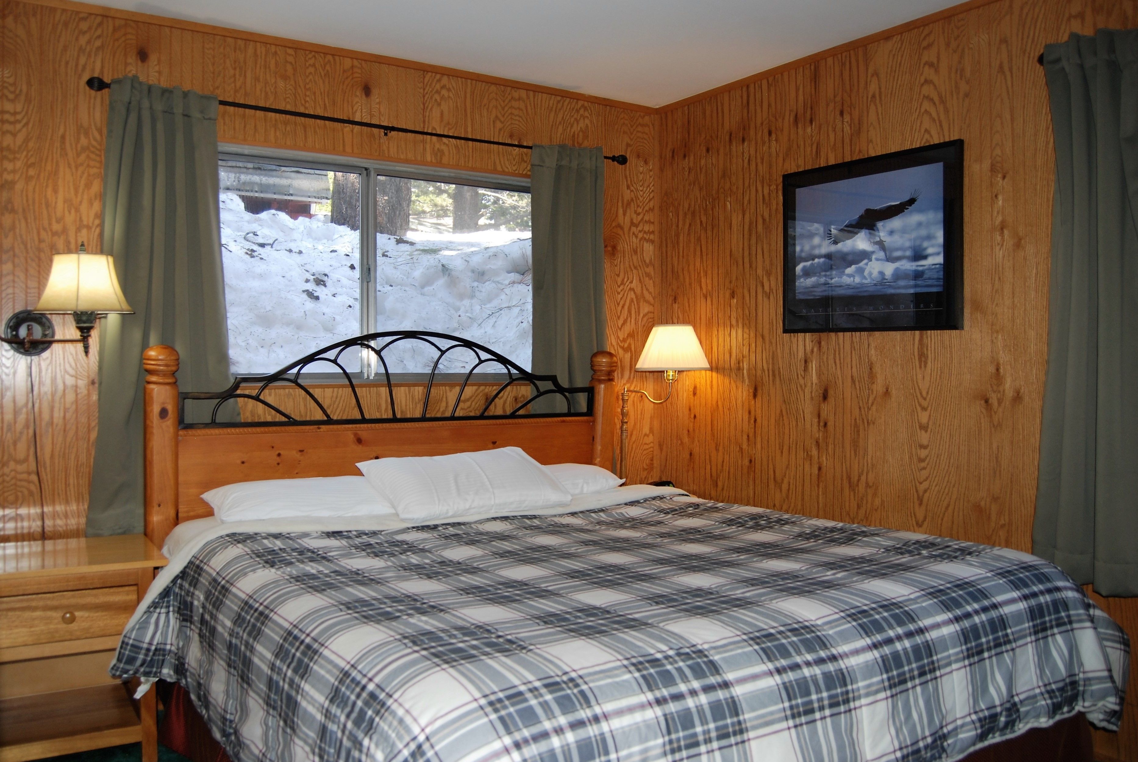 Una habitación revestida de madera con una cama, dos lámparas fijadas a la pared, un ventanal con cortinas y un cuadro enmarcado.