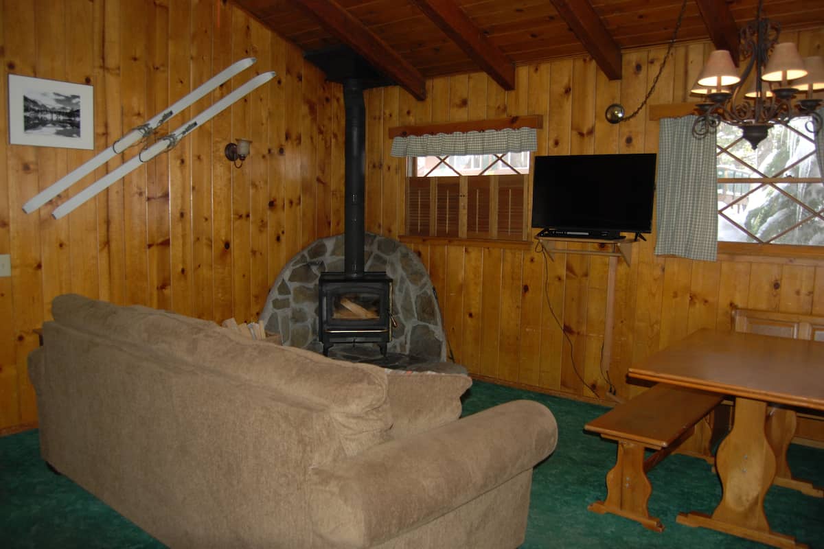 Interior acolhedor de uma cabana com lareira de pedra, um sofá, uma televisão e uma mesa de madeira.