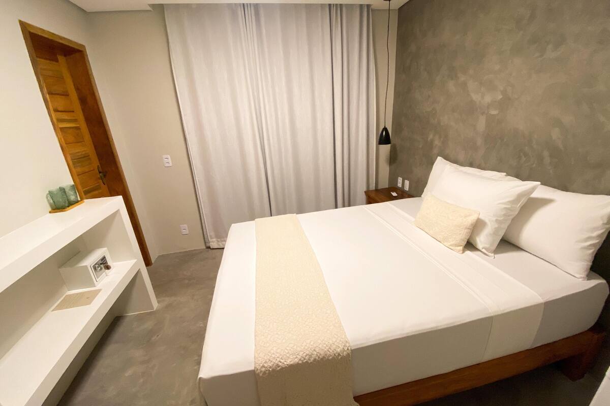 Ein Schlafzimmer mit einem großen Bett, einem Holz-Kopfteil, einem Nachttisch, einer Lampe, einem Fenster mit Vorhängen und einer Holztür.