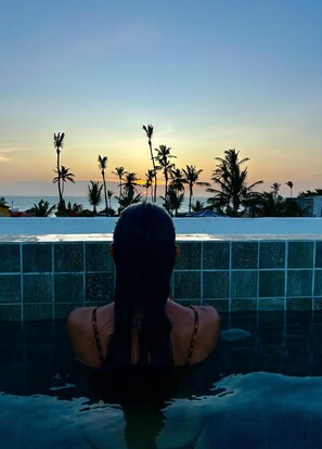 Uma pessoa em uma piscina com vista para palmeiras e o oceano ao pôr do sol.