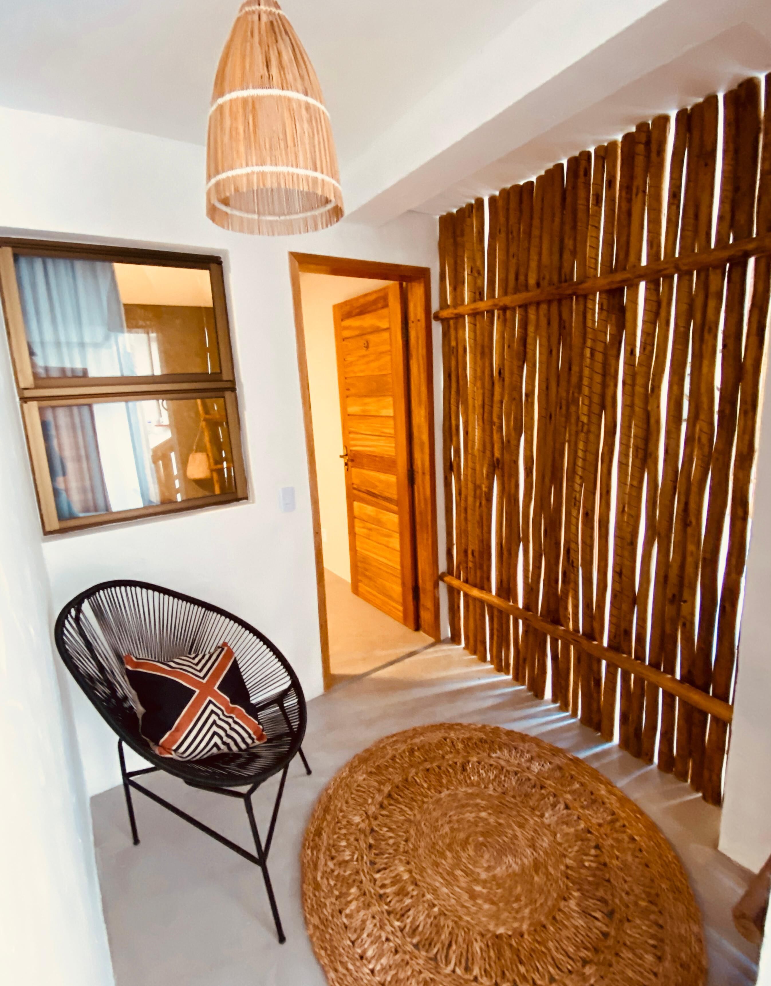 A room with a woven partition, a black chair with a patterned cushion, a woven rug, and a hanging light fixture.
