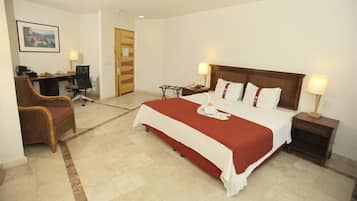 A hotel room with a large bed, two bedside tables with lamps, a desk with a chair, a wicker chair, and a framed picture on the wall.