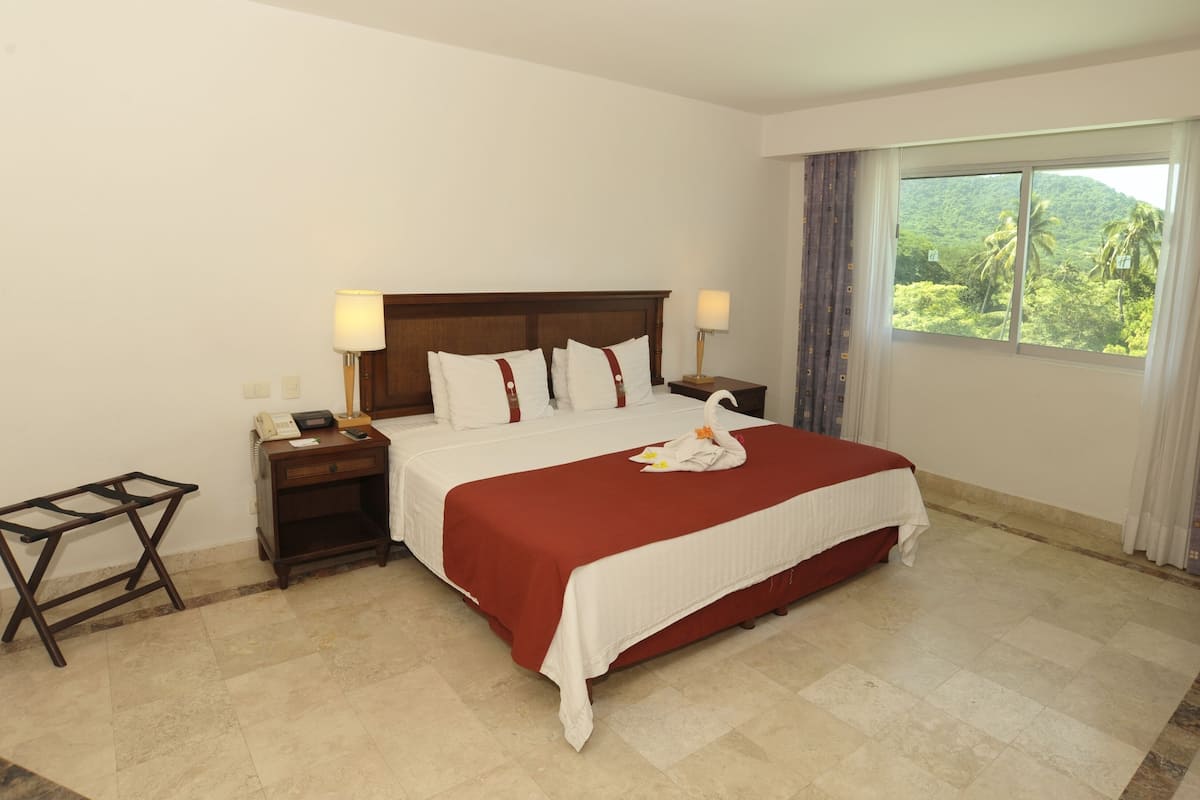 A hotel room with a bed, bedside tables, a nightstand, a window with a view of greenery, and a curtain.