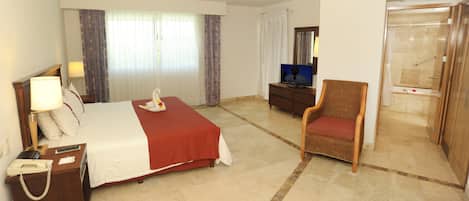 Una habitación de hotel con cama, una silla, escritorio con teléfono, televisor y baño.