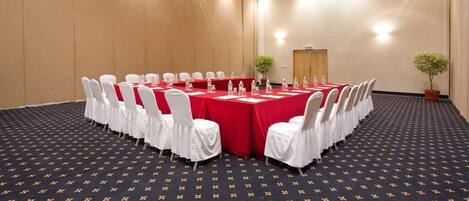 Une salle de conférence avec une longue table, des chaises blanches et une nappe rouge.