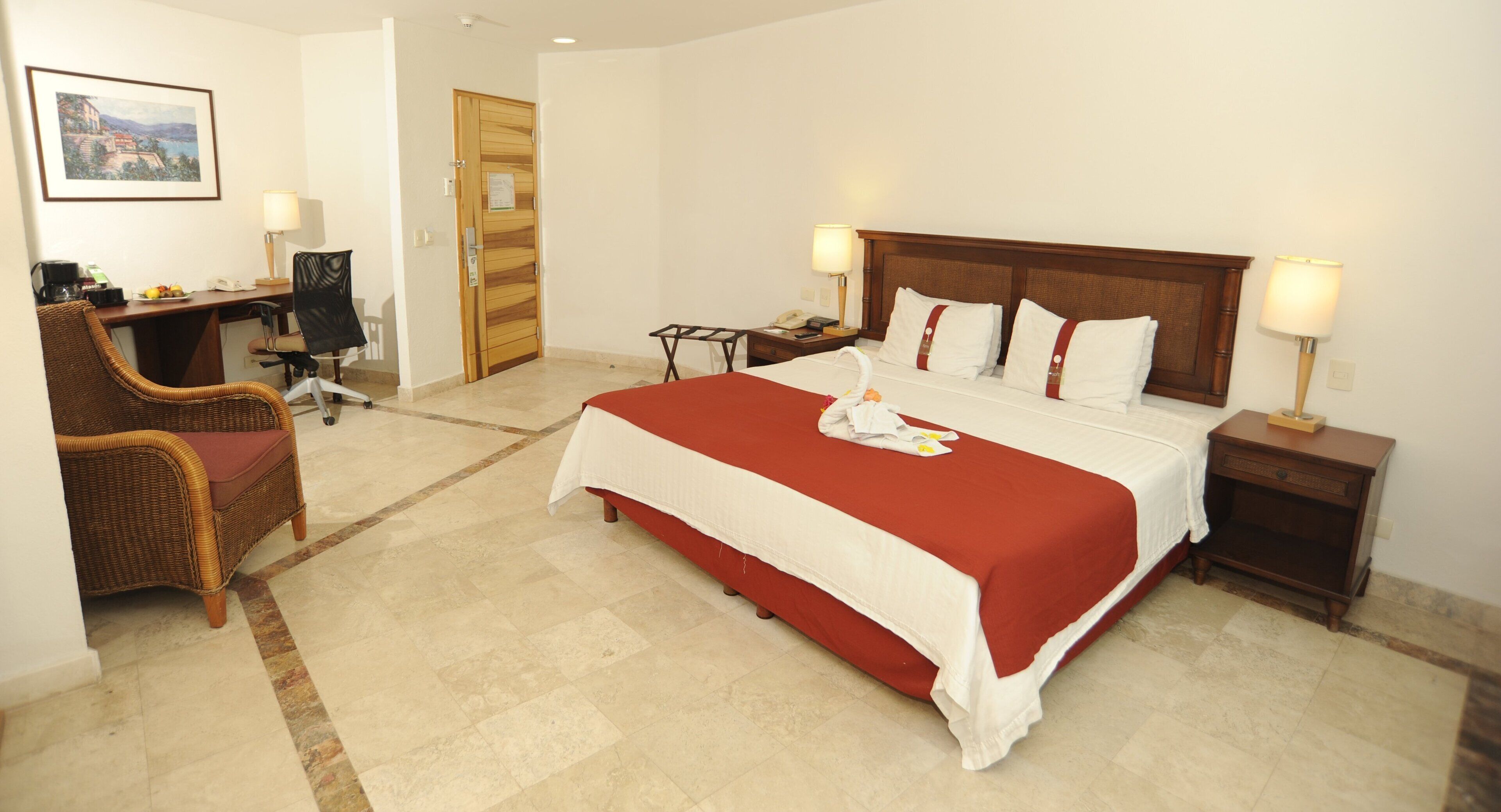 A hotel room with a large bed, a desk with a chair, a wooden door, and a nightstand with a lamp.