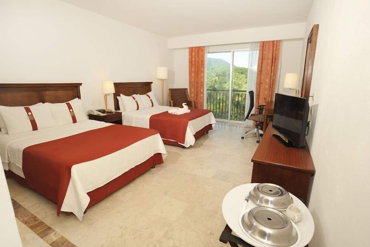 A hotel room with two beds, a TV, a desk with a computer, and a balcony with a view.
