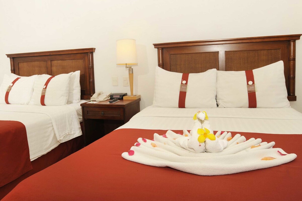 Habitación de hotel con dos camas, cada una con cabecero de madera, ropa de cama blanca y detalles en rojo. Se observa un mesita de noche con una lámpara y un teléfono.
