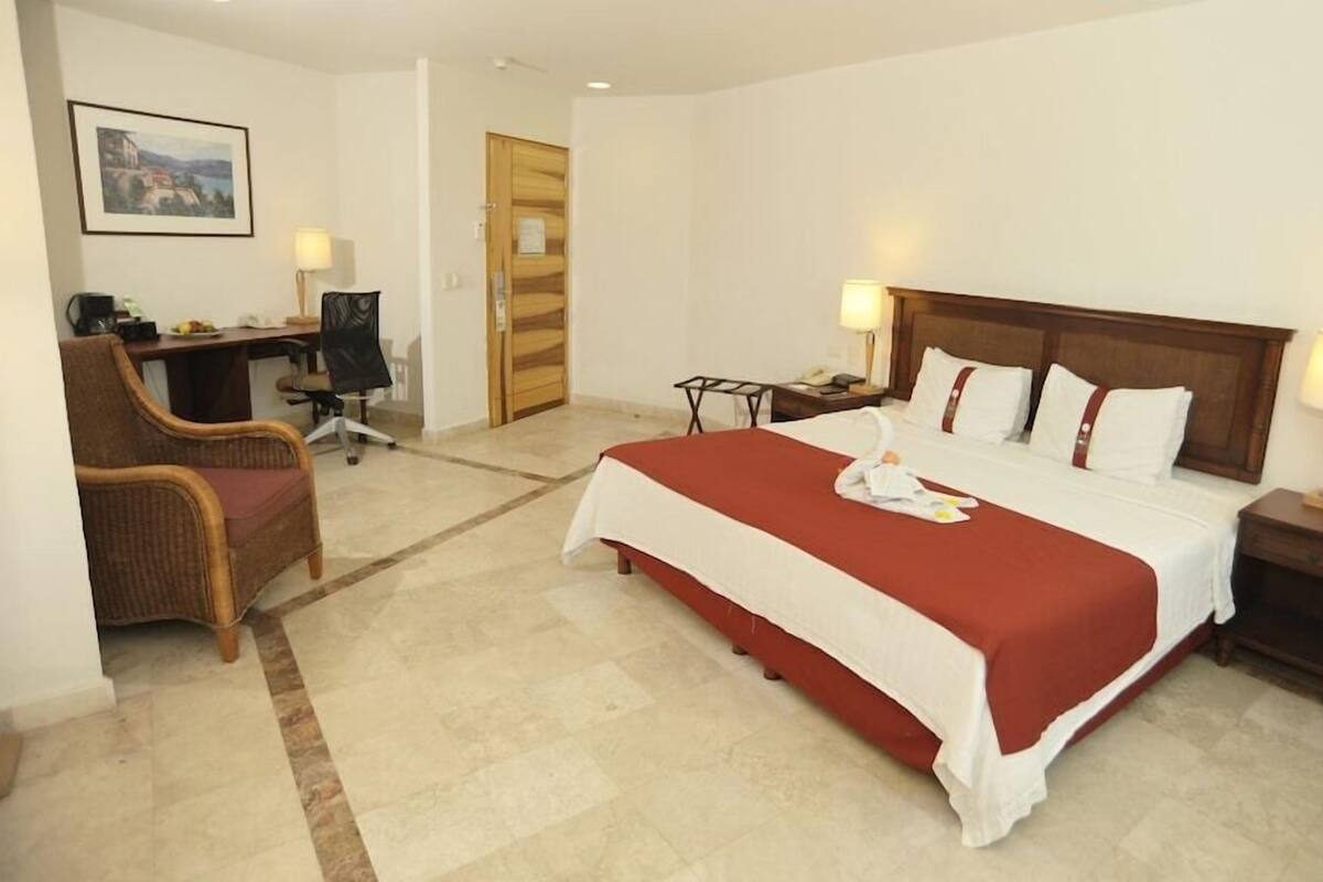 A hotel room with a bed, two bedside tables, a desk with a chair, a lamp, and a framed picture on the wall.