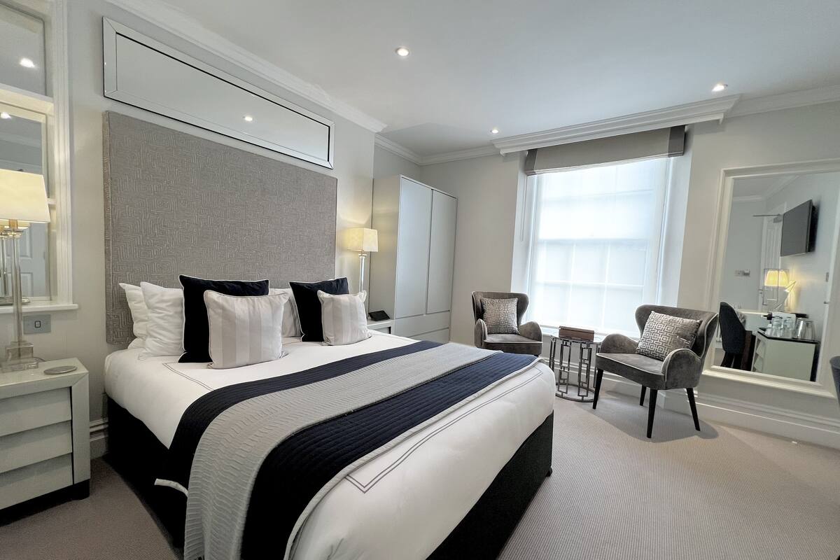 A modern hotel room with a large bed, a sitting area with a chair and a small table, a large mirror, and a window with curtains.