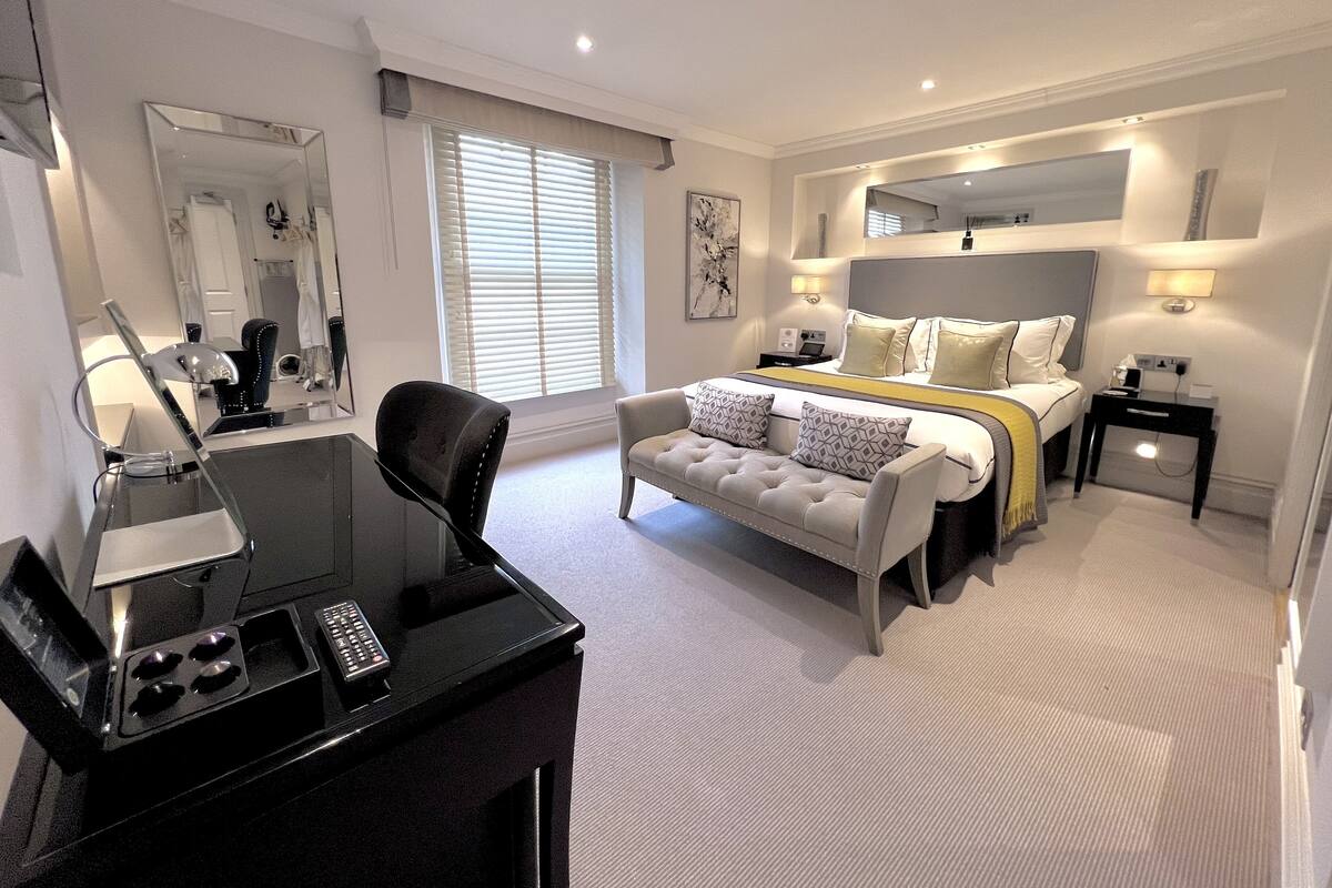 A modern hotel room with a large bed, a sitting area with a sofa, a desk with a computer and a lamp, and a large mirror.