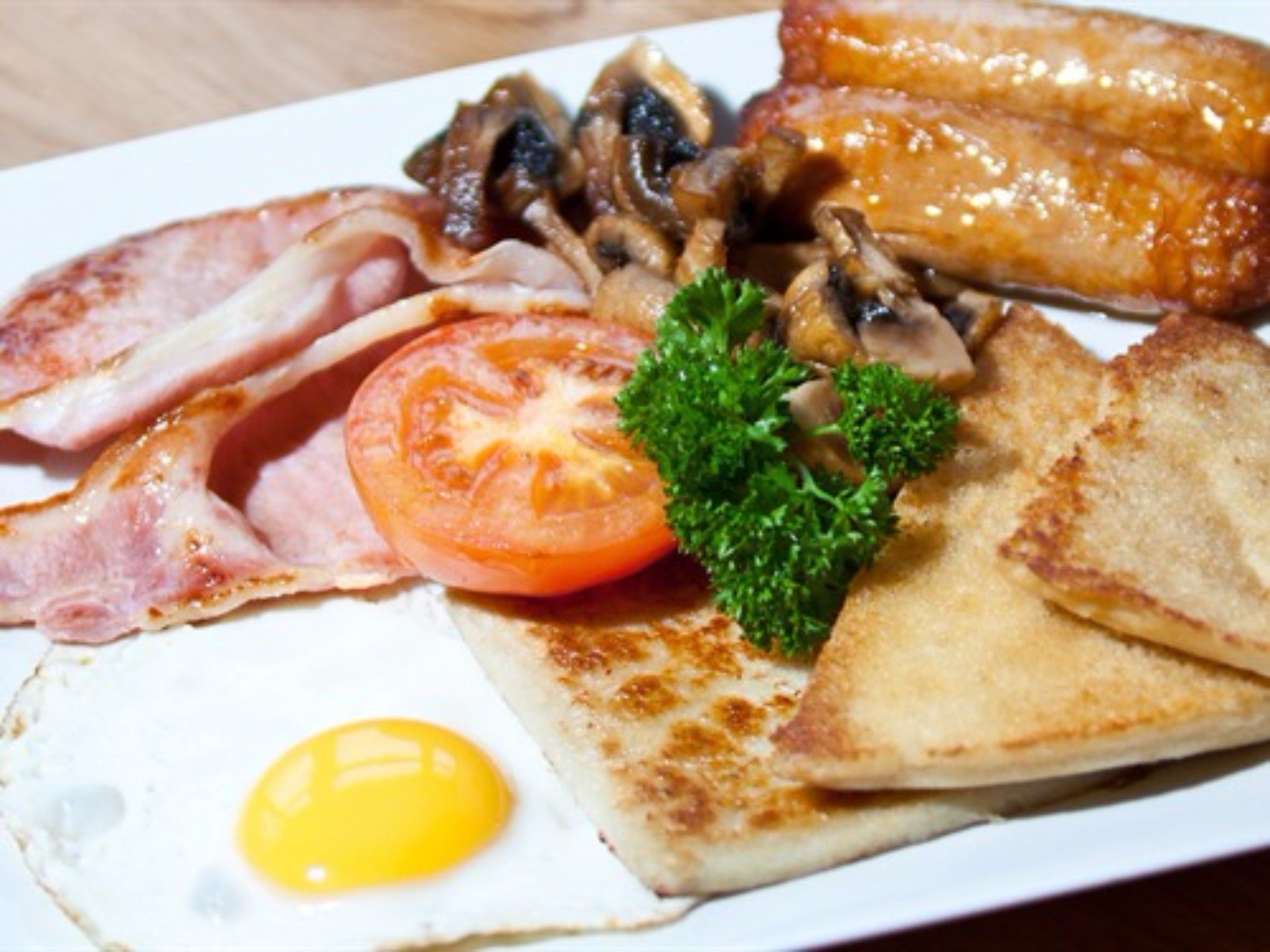 A plate of breakfast food including bacon, sausages, mushrooms, a fried egg, toast, and a tomato.