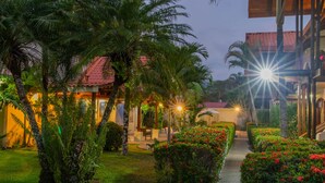 Un sendero bien iluminado que conduce a un edificio con techo rojo, rodeado de exuberante vegetación y plantas con flores.