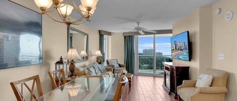 A spacious living room with a large glass dining table, comfortable seating, a chandelier, and a cityscape painting.