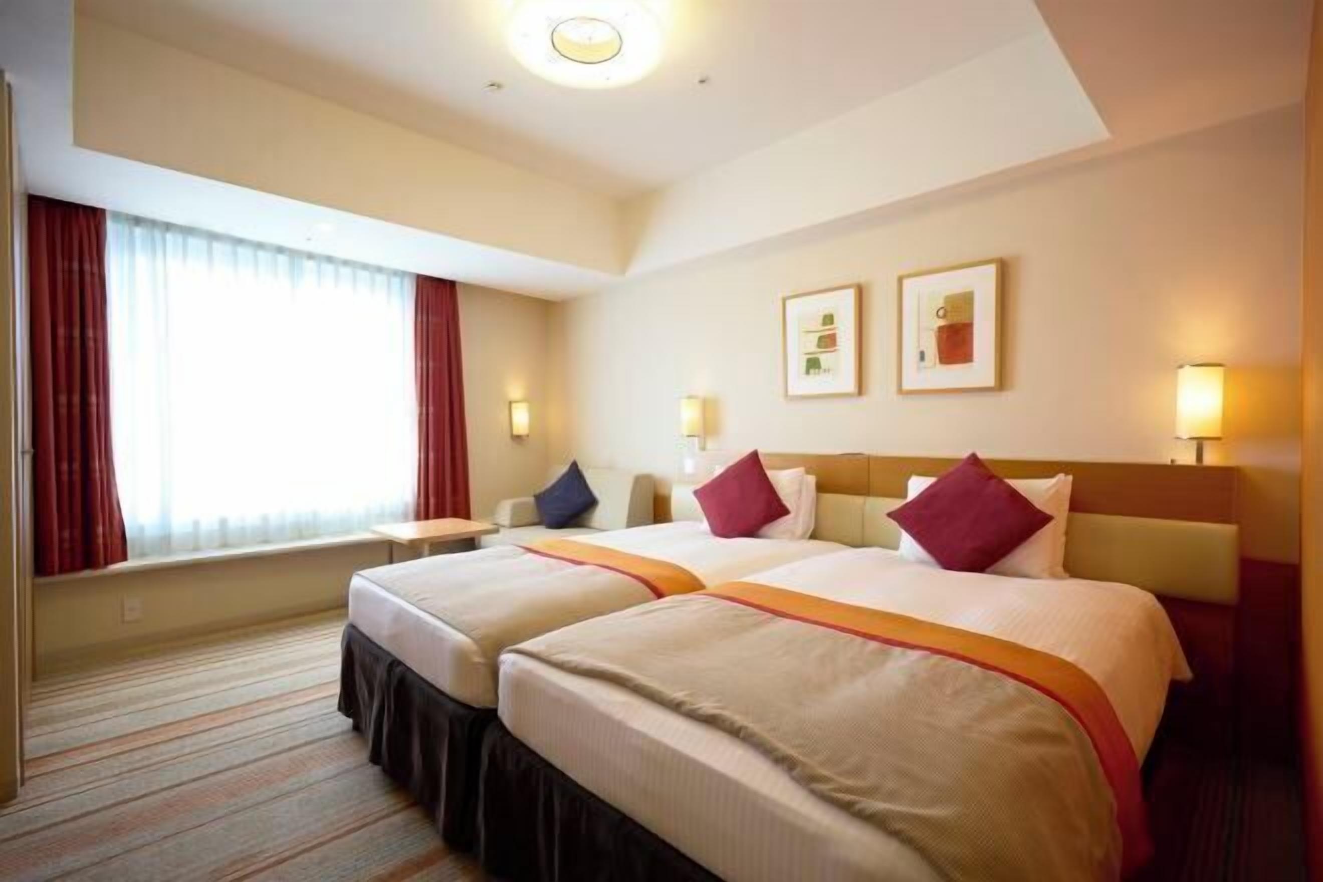 Ein Hotelzimmer mit zwei Betten, einem Fenster mit Vorhängen, einem Nachttisch und einer Wandlampe.