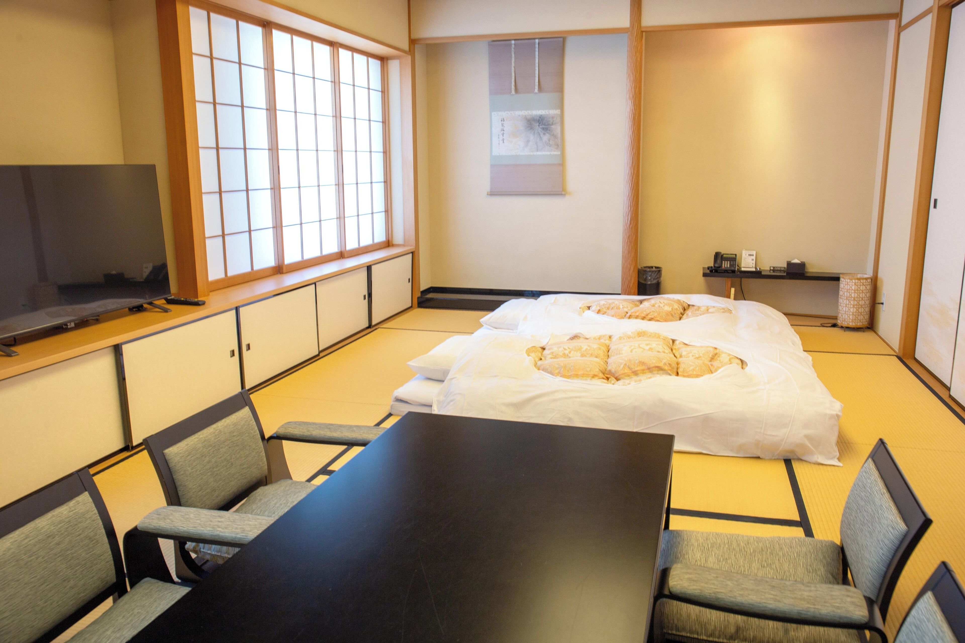 A traditional Japanese-style room with tatami flooring, a low table with chairs, a bed with futon bedding, and a television on a low stand.