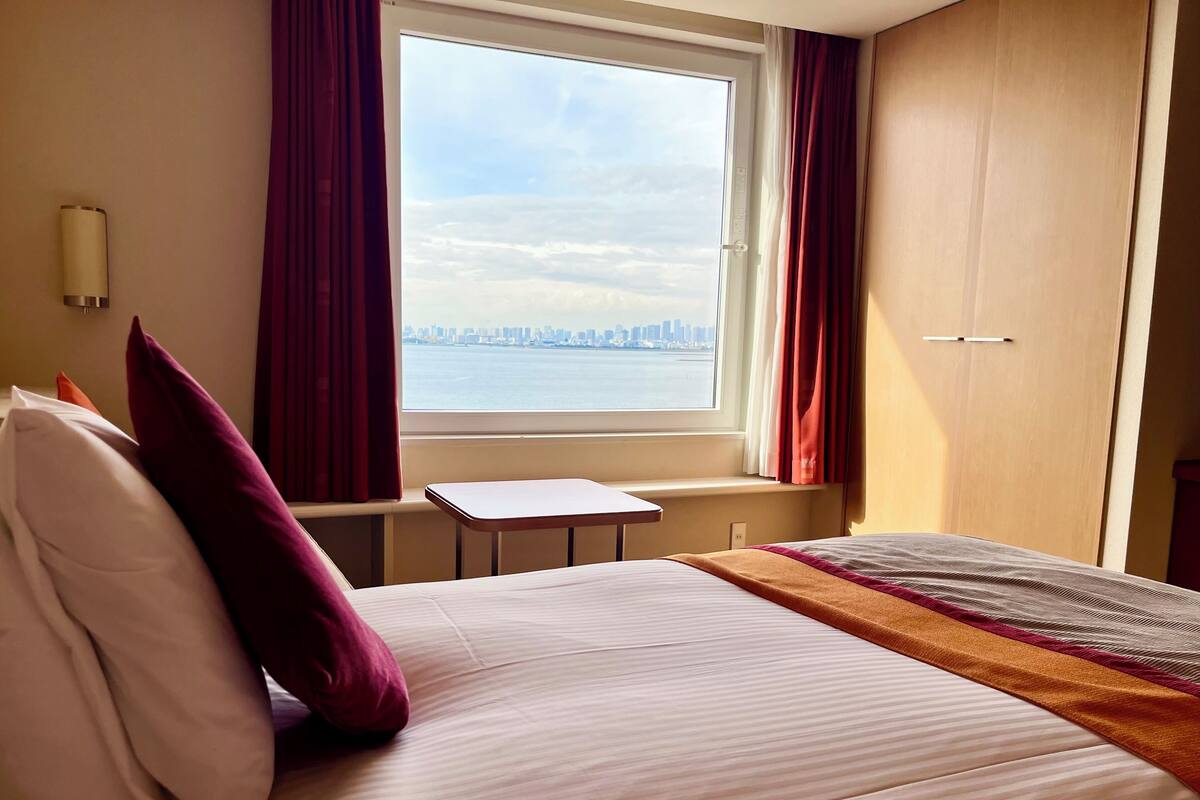 A hotel room with a bed, a small table, a view of a city skyline through a window, and a door.